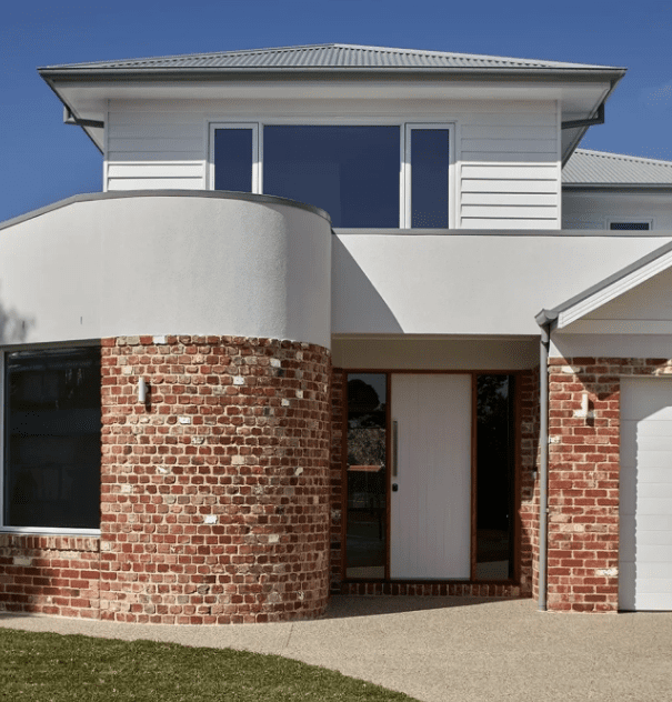 Modern two-story house with white siding and red brick accents, large windows, a wooden front door, and an attached garage.
