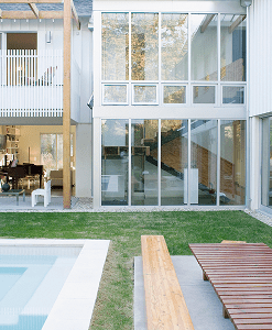 Modern house with large glass windows, a wooden bench, and a pool in a grassy backyard. Indoor furniture and open shelving visible through the windows.