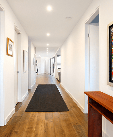A modern hallway with white walls, wooden flooring, a black runner rug, and several doors along each side. A framed artwork and a wooden console table are visible on the right.