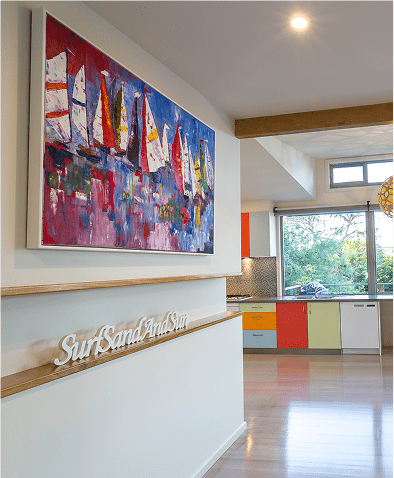 Modern kitchen with colorful cabinets, a large window, and a wall displaying an abstract sailboat painting and a "Surf Sand And Sun" decorative sign.