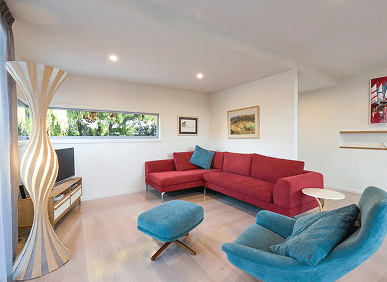 Modern living room with a red sectional sofa, teal armchair and ottoman, unique floor lamp, TV, and wall art, featuring light wood flooring and white walls.