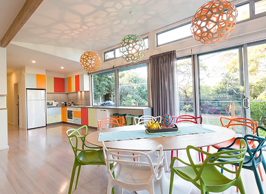 A modern kitchen and dining area with colorful cabinets, mismatched chairs, large windows, pendant lights, and a round table set with fruit.