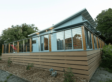 Modern single-story house with large windows, a slanted roof, and a wooden deck enclosed by wire and wood railing; surrounded by trees and mulch landscaping.