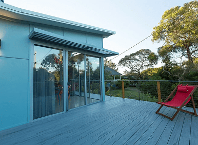 A modern patio with wooden decking, a glass sliding door, a retractable awning, and a red lounge chair, overlooking a green yard with trees.