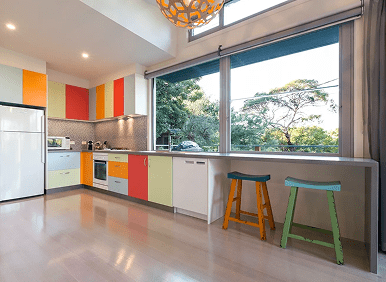 Modern kitchen with colorful cabinets, large windows, two stools at the counter, and appliances including a refrigerator, oven, and microwave.